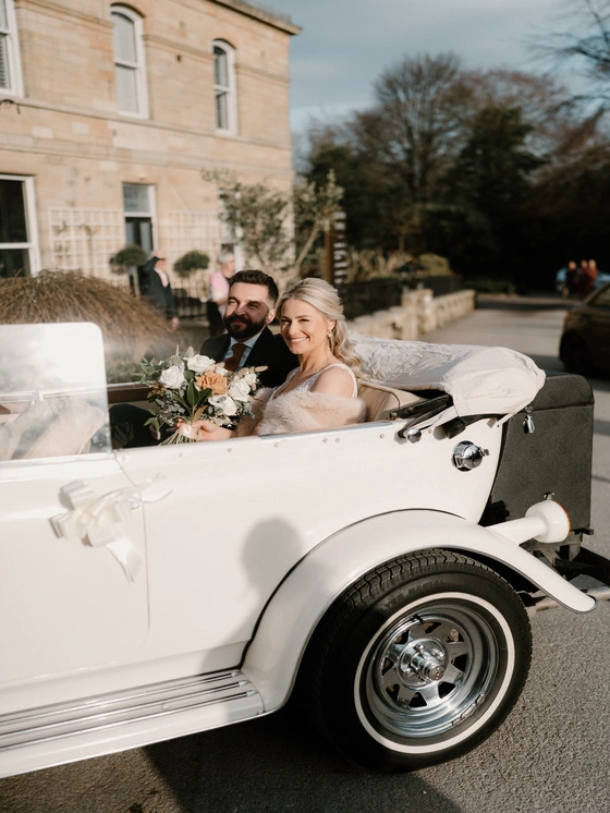 Bride and groom in a classic convertible car outside the entrance of Manor House Lindley