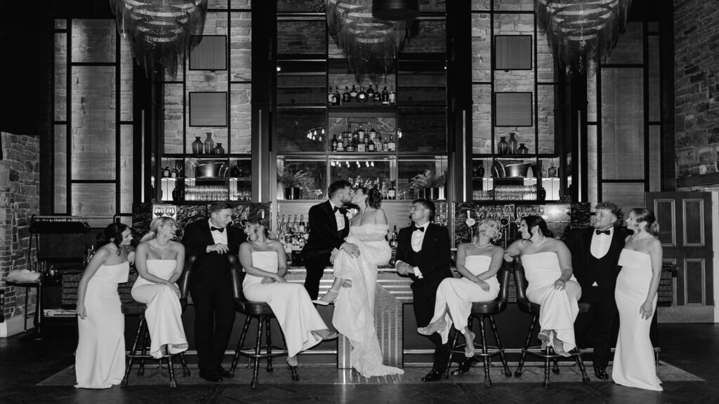 Black and white picture of bride and groom kissing at the bar surrounded by family and friends.