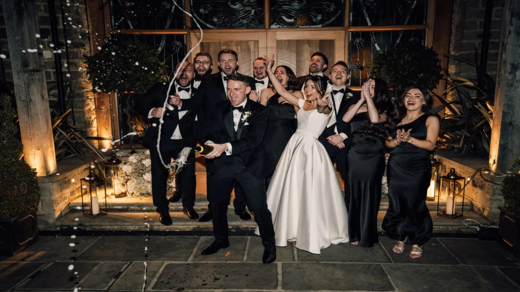 Bride and groom spraying bottle of Champagne surrounded by bridesmaid, best man, and friends